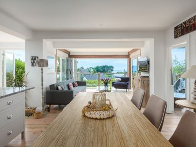 A dining table and chairs in an open living area with a sofa, armchair, and a view of houses and trees at Trewartha in Praa Sands