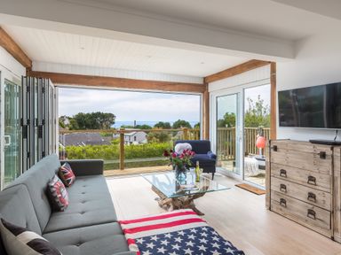 A living room with a gray sofa and American flag blanket by a glass table with flowers and a wooden chest at Trewartha in Praa Sands