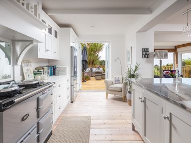 A kitchen with white cabinets a gray oven and an island with a marble countertop leading to a wooden deck with outdoor furniture at Trewartha in Praa Sands