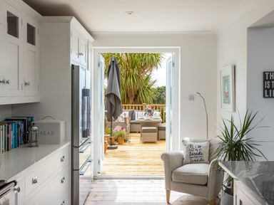 A kitchen with white cabinets a refrigerator and an armchair with a cushion next to a door opening to a wooden deck with outdoor seating at Trewartha in Praa Sands