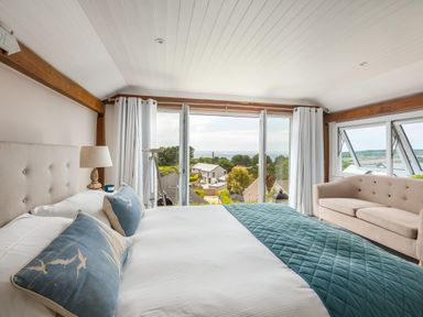 A bedroom with a bed with blue pillows and quilt a beige sofa and large windows showing a view of houses and trees at Trewartha in Praa Sands
