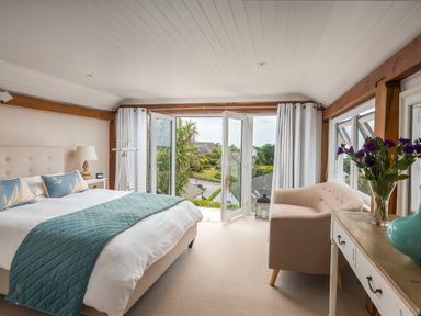 A bedroom with a bed with blue pillows and quilt near open balcony doors and a small armchair at Trewartha in Praa Sands