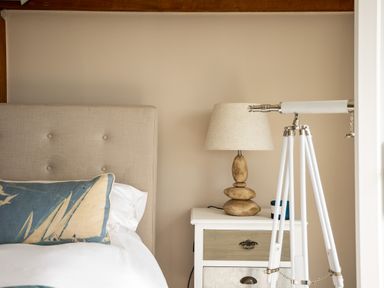 A bedroom with a beige headboard a blue pillow a bedside table with a stone lamp and a white telescope at Trewartha in Praa Sands