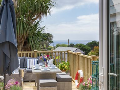 An outdoor dining area with a table set for six on a wooden deck overlooking houses and the sea at Trewartha in Praa Sands