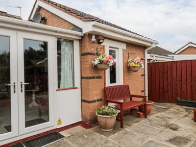 An outdoor patio area with a red wooden bench two hanging flower baskets and potted plants near a brick house at Tartaria in Widdrington