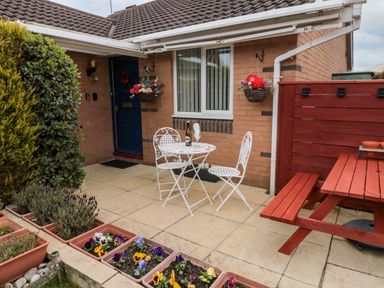 A patio with a white metal table and chairs a bottle on the table flower pots and a red wooden picnic table at Tartaria in Widdrington