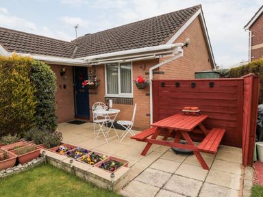 An outdoor patio area with a small white table and chairs set and a red wooden picnic table with flowers in pots and planters at Tartaria in Widdrington