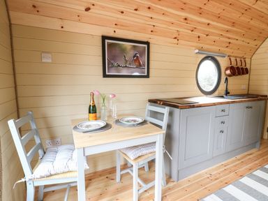 A small dining area with a table set for two and a kitchen counter with a sink and hanging pans at Willow Pod in Arundel West Sussex