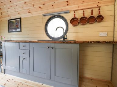 A kitchen with grey cabinets a round window above the sink copper pans hanging on the wall and wooden walls at Willow Pod in Arundel West Sussex