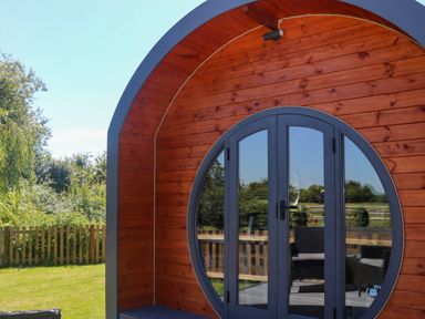 An outdoor wooden pod with circular glass doors and garden furniture in the background at Willow Pod in Arundel West Sussex