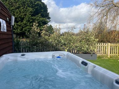 An outdoor hot tub with bubbling water next to a wooden shed and plants with a picket fence and trees in the background at Willow Pod in Arundel West Sussex