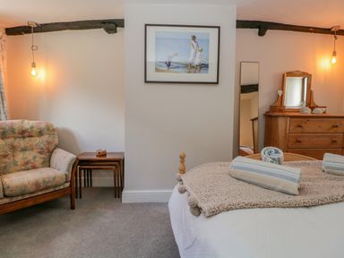 A bedroom with a bed, folded towels, a dresser with mirror, an armchair, nesting tables, and a framed picture on the wall at Ashness in Bassenthwaite near Keswick