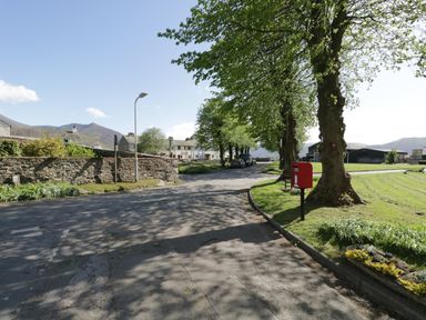 A street with trees lining the side and a red postbox near a grassy area in a rural setting at Ashness in Bassenthwaite near Keswick