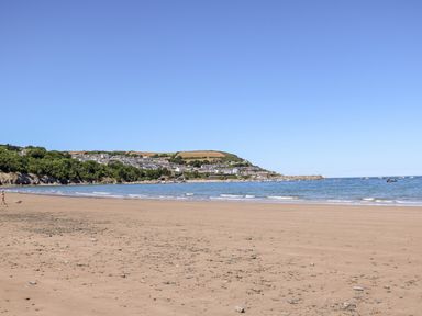 A sandy beach with scattered stones people and sea with houses and trees in the background at Cwtsh Cragen in New Quay