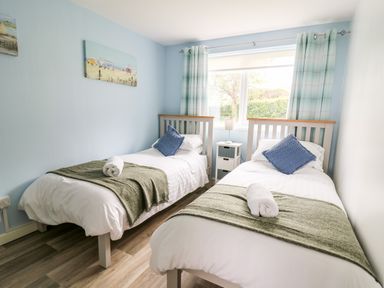A bedroom with two single beds with green throws and blue pillows a nightstand with a lamp and a window with curtains at Cwtsh Cragen in New Quay
