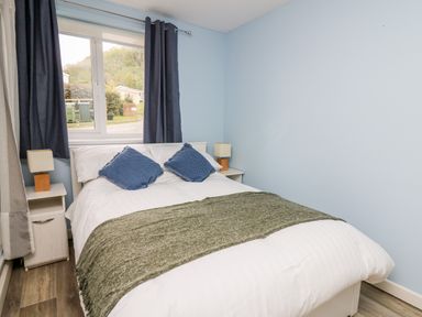 A bedroom with a double bed, blue pillows, bedside tables with lamps, and a window with curtains at Cwtsh Cragen in New Quay