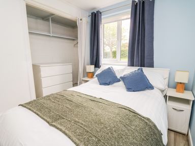 A bedroom with a double bed with pillows and a blanket two bedside tables with lamps a chest of drawers and an open closet at Cwtsh Cragen in New Quay