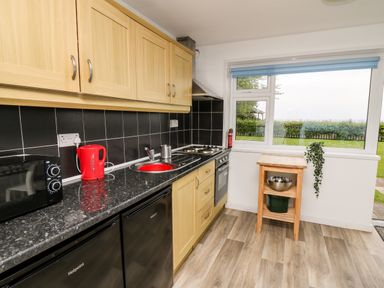 A kitchen with wooden cabinets black countertop a red kettle a microwave a stove and a small wooden table under a window at Cwtsh Cragen in New Quay