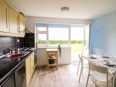 A kitchen with wooden cabinets black countertop dining table with white chairs and garden view at Cwtsh Cragen in New Quay