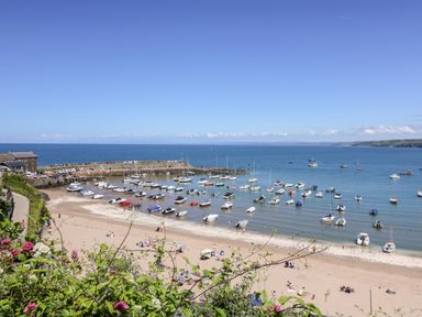 A beach with people on the sand and boats anchored near a stone pier at Cwtsh Cragen in New Quay
