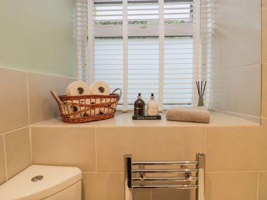 A bathroom window sill with a basket of toilet rolls soap dispensers a towel and a reed diffuser at 1 Tower House in Cartmel