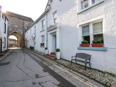 A narrow street with white buildings flower pots on windows and a bench near the entrance at 1 Tower House in Cartmel