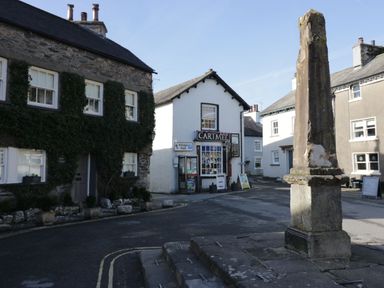 A street scene with stone and white buildings and a tall stone monument in the center at 1 Tower House in Cartmel