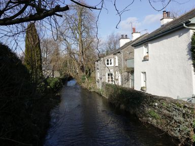 A river flowing beside stone and white houses with trees and plants at 1 Tower House in Cartmel