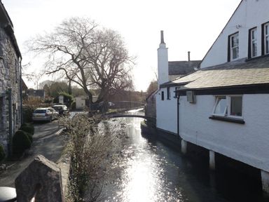 A stone wall beside a narrow river with houses on one side and a small iron bridge over the river at 1 Tower House in Cartmel
