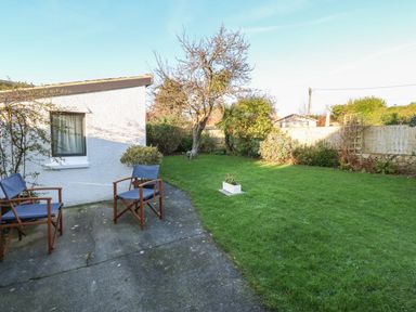 A garden with two wooden chairs on a concrete patio next to a white building and a grassy lawn with trees at Disgwylfa in Moelfre