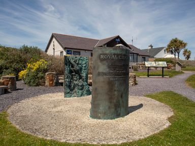 An outdoor memorial with a curved pillar and a relief sculpture near a building with lifeboats signs at Disgwylfa in Moelfre