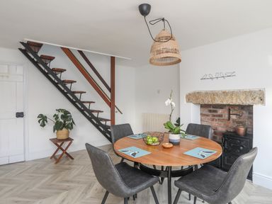 A dining area with a round wooden table set for four, gray chairs, a staircase, a brick fireplace with a wood stove, and a plant on a stand at Disgwylfa in Moelfre