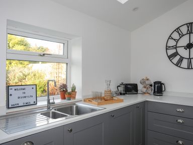 A kitchen corner with a double sink under a window and countertops holding a tray with glasses a carafe a toaster a kettle and a mug holder at Disgwylfa Moelfre