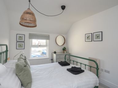 A bedroom with a green metal bed frame, pillows, folded towels, wall art, a window, a dresser with a mirror, and a pendant light at Disgwylfa in Moelfre
