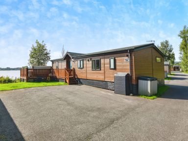 A wooden lodge with a small deck and steps with trees and water in the background at Gerrida Lodge South Lakeland Leisure Village near Tewitfield