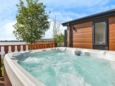 An outdoor hot tub on a wooden deck next to a cabin with a window and trees by a lake at Gerrida Lodge South Lakeland Leisure Village near Tewitfield