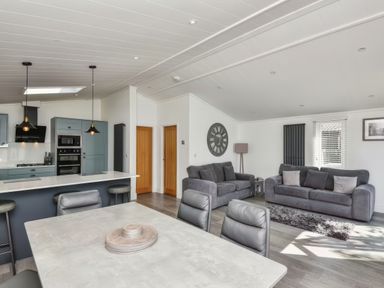 An open-plan living area with a kitchen, dining table, two sofas, wall clock, and a floor lamp at Gerrida Lodge in South Lakeland Leisure Village near Tewitfield