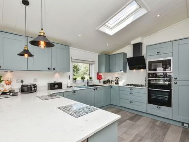 A kitchen with blue cabinets a gas stove built-in oven hanging pendant lights and a window at Gerrida Lodge South Lakeland Leisure Village near Tewitfield