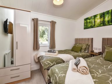 A bedroom with two single beds a wardrobe a wall-mounted TV and a window with curtains at Gerrida Lodge in South Lakeland Leisure Village near Tewitfield