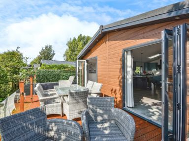 An outdoor deck with grey wicker chairs and a glass table by a wooden cabin with open doors to the kitchen at Gerrida Lodge South Lakeland Leisure Village near Tewitfield