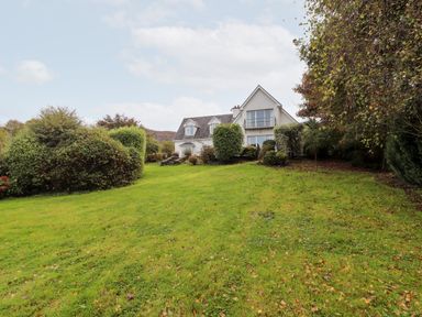 A house with a sloped roof and balcony surrounded by bushes and trees with a grassy lawn in front at Seascape Lodge in Kenmare County Kerry