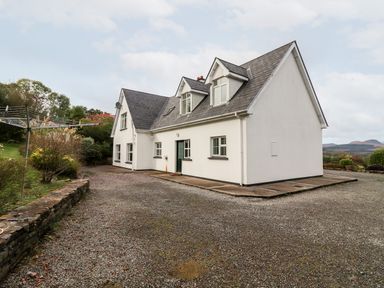 A white two-story house with a sloped roof and windows on a gravel driveway with surrounding greenery at Seascape Lodge in Kenmare County Kerry