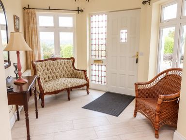 An entryway with a floral upholstered wooden settee a wicker chair a wooden table with a lamp and a white door with stained glass panels at Seascape Lodge in Kenmare County Kerry