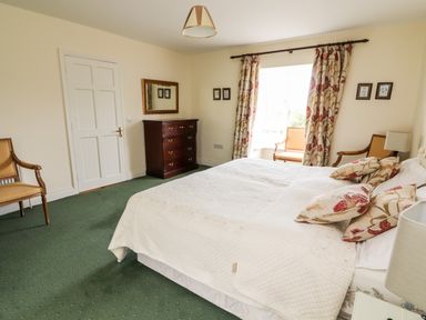 A bedroom with a bed a wooden dresser two chairs a mirror and floral curtains at Seascape Lodge in Kenmare County Kerry