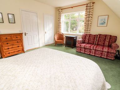A bedroom with a bed covered by a white quilt a wooden dresser two white doors a window with patterned curtains a wooden chair a side table and a red patterned sofa at Seascape Lodge in Kenmare County Kerry