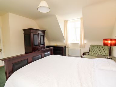 A bedroom with a bed covered with a white blanket an armchair a wooden wardrobe and a window with curtains at Seascape Lodge in Kenmare County Kerry