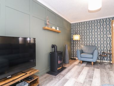 A living room with a television on a wooden stand a black stove a blue armchair and patterned wallpaper at Alfresco Lodge Skiptory Howe 37B White Cross Bay near Windermere