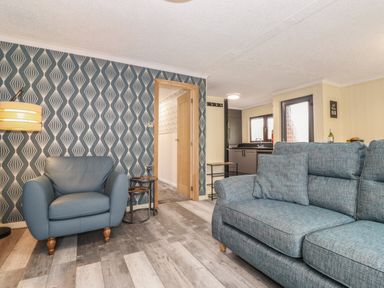 A living room with a blue armchair a blue sofa patterned wallpaper and a view into a kitchen at Alfresco Lodge Skiptory Howe 37B White Cross Bay near Windermere