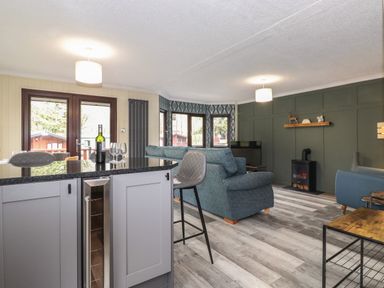 An open living room and kitchen area with bar stools wine bottle and glasses a blue sofa TV and electric fireplace at Alfresco Lodge in Skiptory Howe 37B White Cross Bay near Windermere