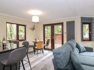 A living area with a blue sofa a dining table with chairs bar stools and glass doors to a wooden deck at Alfresco Lodge in Skiptory Howe 37B White Cross Bay near Windermere